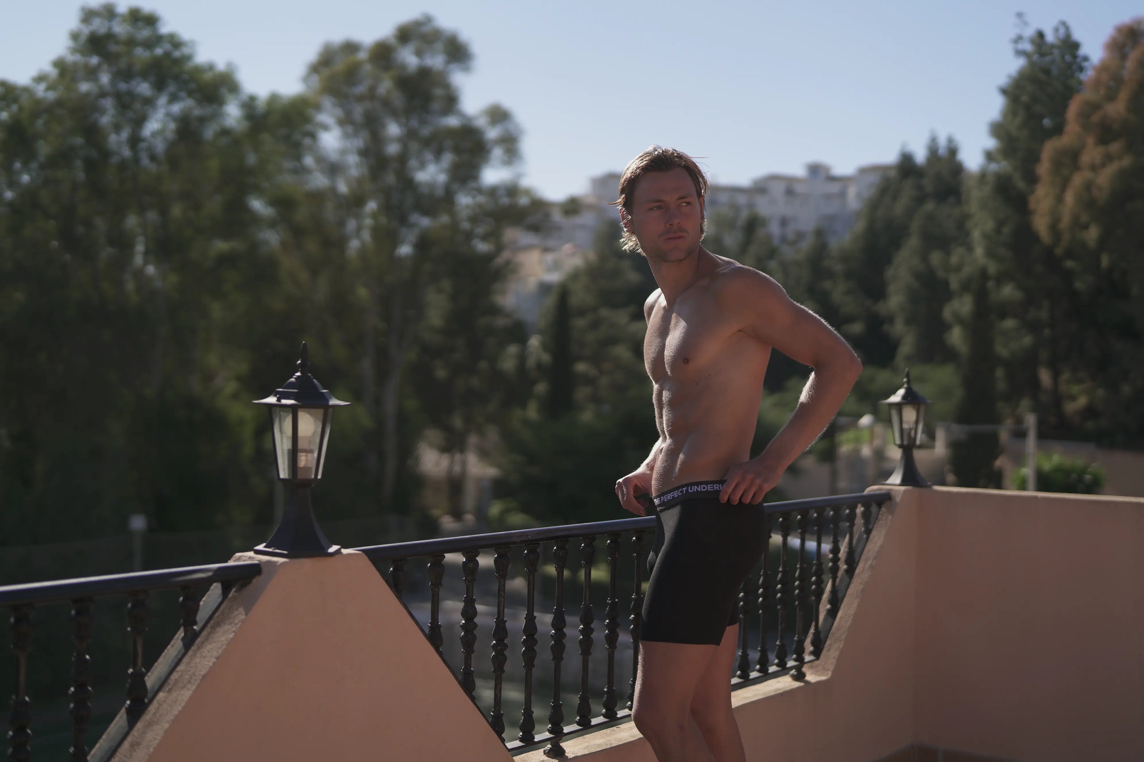 Man standing in boxer briefs on a balcony with trees and buildings in the background