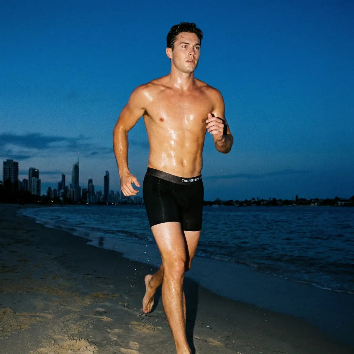 The Perfect Underwear bamboo boxer briefs in black, shown worn on a male model running along a beach with a city skyline in the background at dusk.